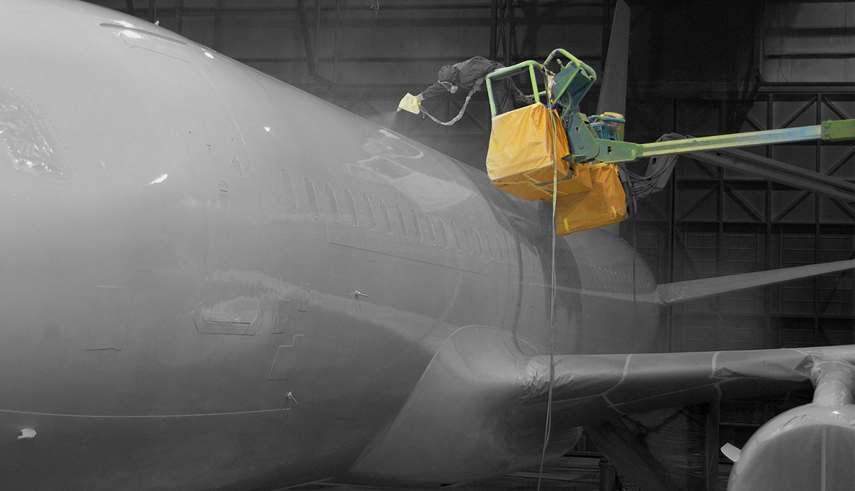 A man in a spray suit standing on a cherry picker sprays the body of an aeroplane. The picker has yellow protective covers around it's sides and base, while the mans spray gun has a white protective spray gun cover.